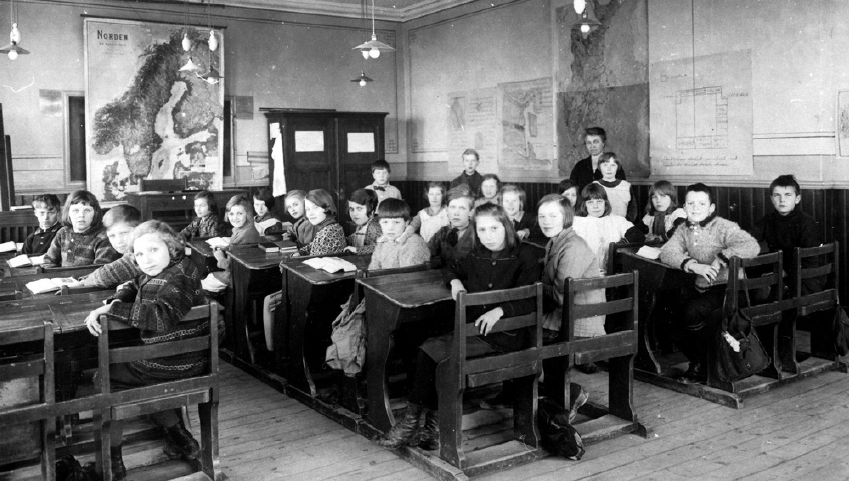 En äldre svartvit bild med en skolklass som sitter vid sina bänkar i en sal.