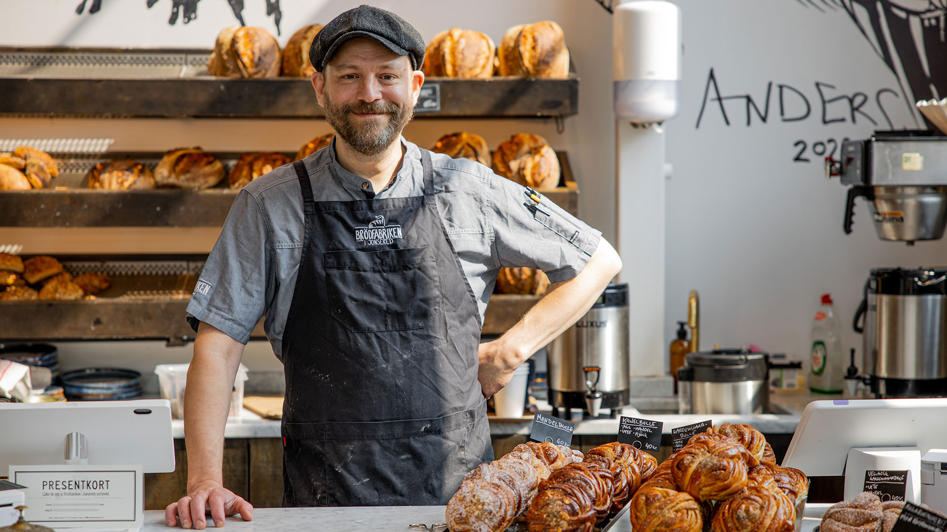 Bild på person i keps och förkläde som står bakom affärsdisk i butiksmiljö med bullar och bröd.