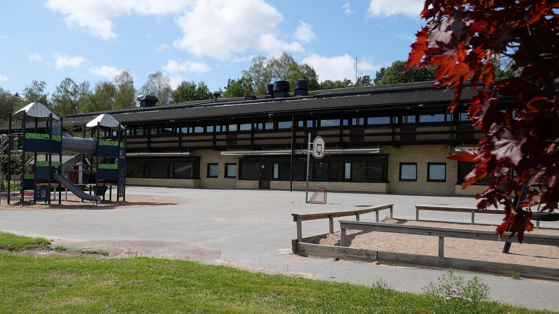 Skolbyggnaden i två våningar syns i bakgrunden, med skolgården framför. På skolgården framför skolbyggnaden syns en klätterborg, basketkorg och sandlåda. 