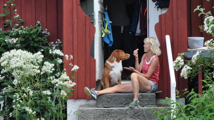 Bild på en skrattande kvinna och hennes hund som sitter tillsammans på trappan till huset med öppen ytterdörr.