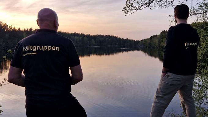 Bilden visar två män. Männen står och tittar mot en sjö. Männen har ryggen vänd mot kameran. På deras ryggar står det Fältgruppen. 