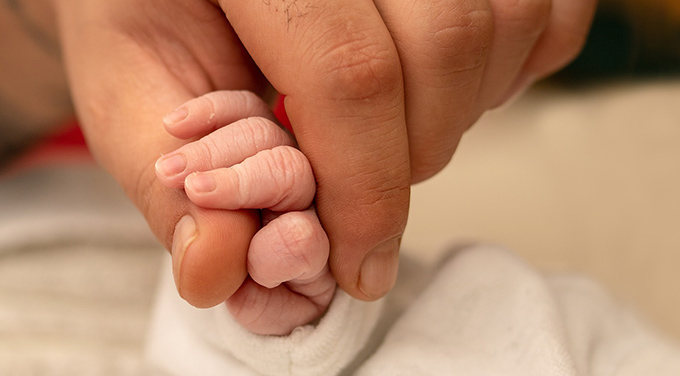 Bilden visar en nyfödd bebis lilla hand som kramar om en vuxen persons finger.