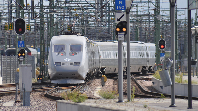 En bild på ett grått SJ-tåg som är på väg in till en perrong. I bakgrunden syns stolpar, elledningar och ljussignaler. 