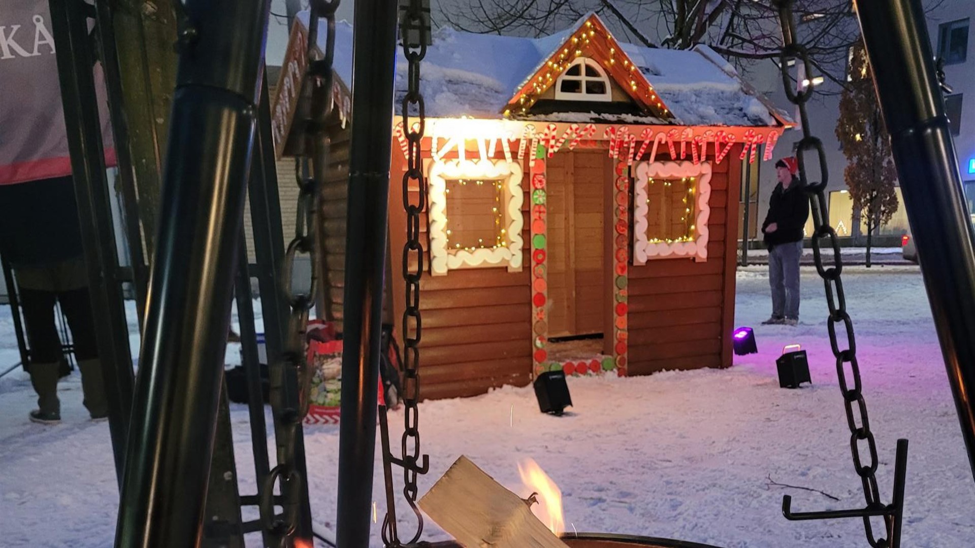 Bild på ett pepparkakshus i mänsklig storlek framför en brasa. Snö ligger på marken och i bakgrunden syns en ung kille iklädd tomteluva.