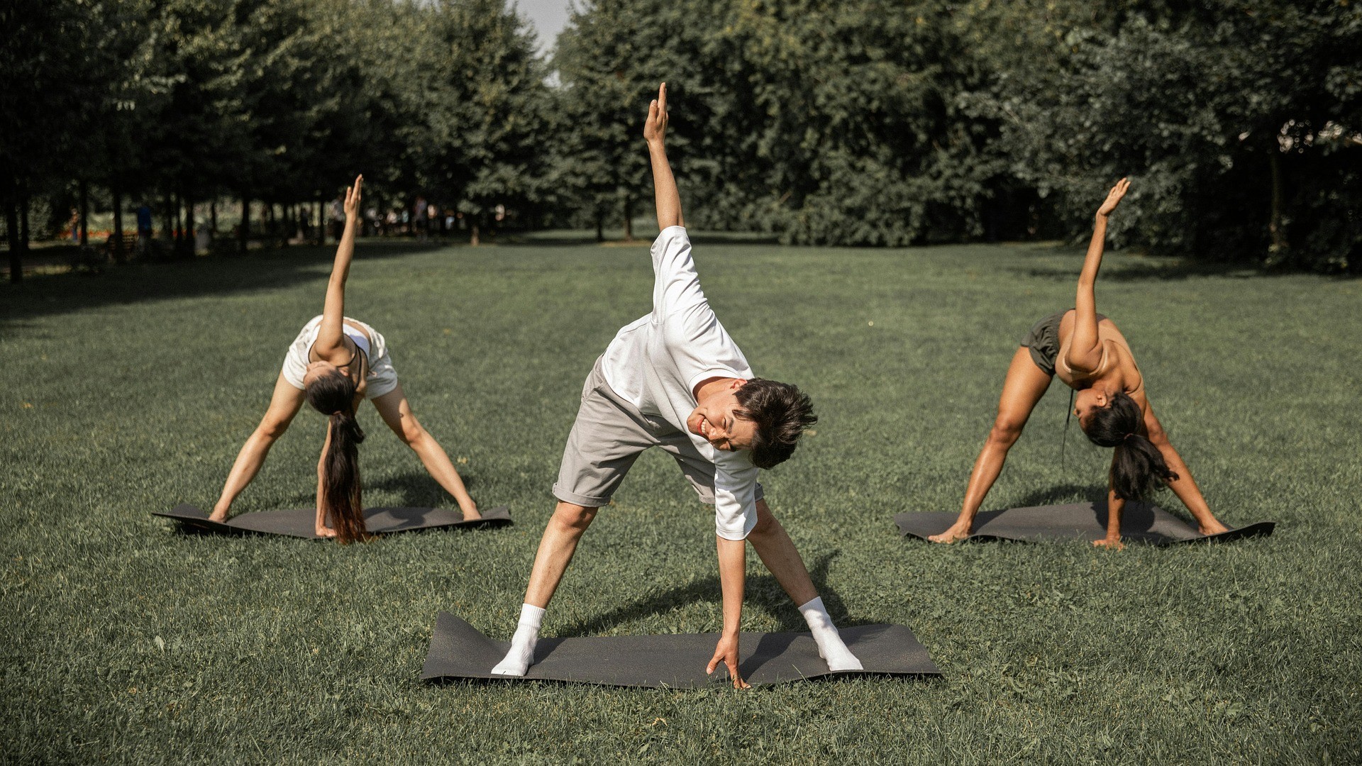 Bild på tre personer som utövar stretching på varsin yogamatta ute i naturen.